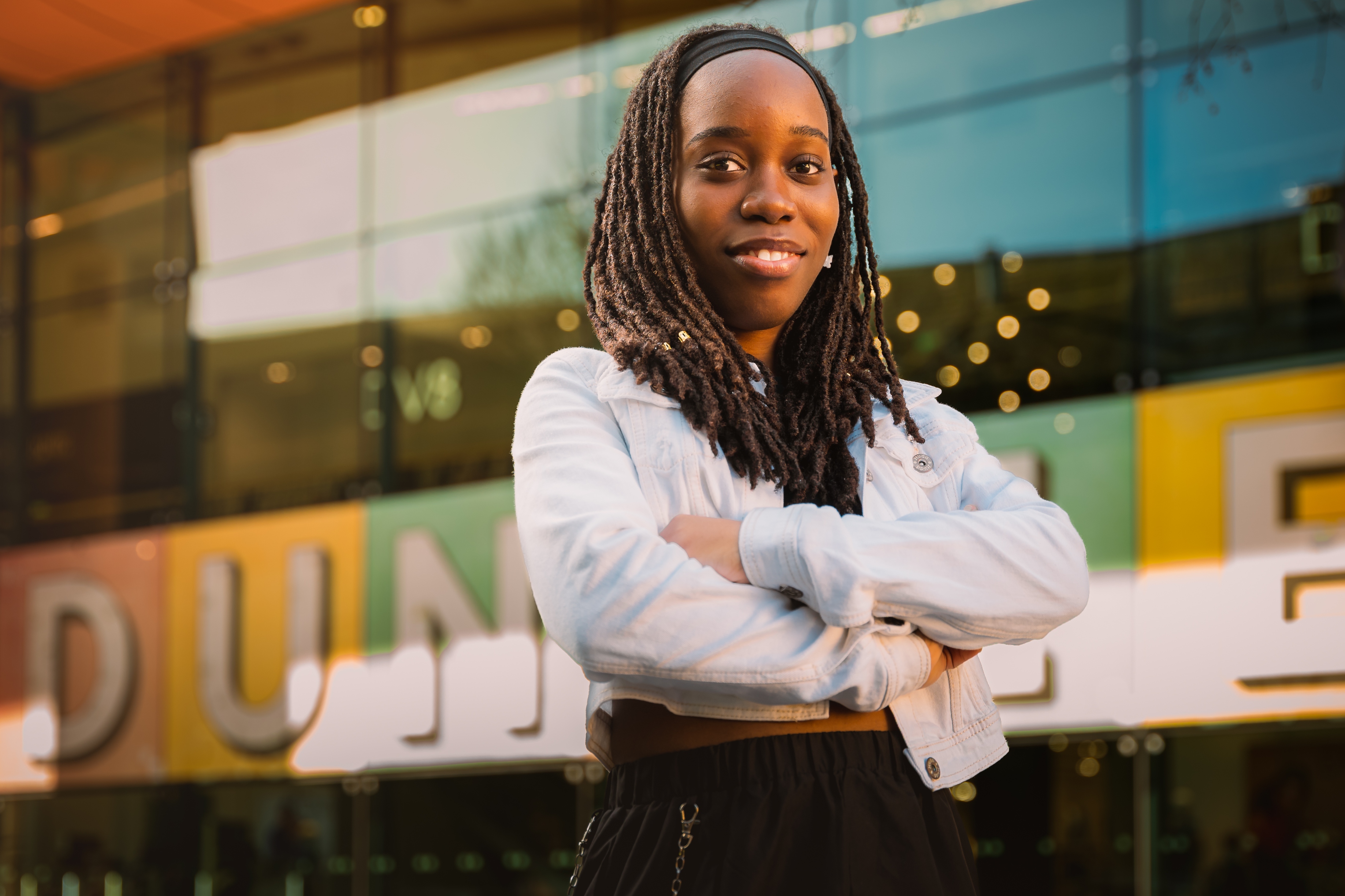 A woman standing with her arms crossed in front of her she smiles at the camera, she stands outside with a window in the background and writing in big letters that spells out 'Dundee'. 