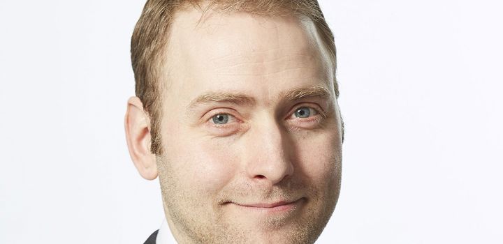 A headshot of a man against a white background, he has short light hair and smiles facing the camera. 