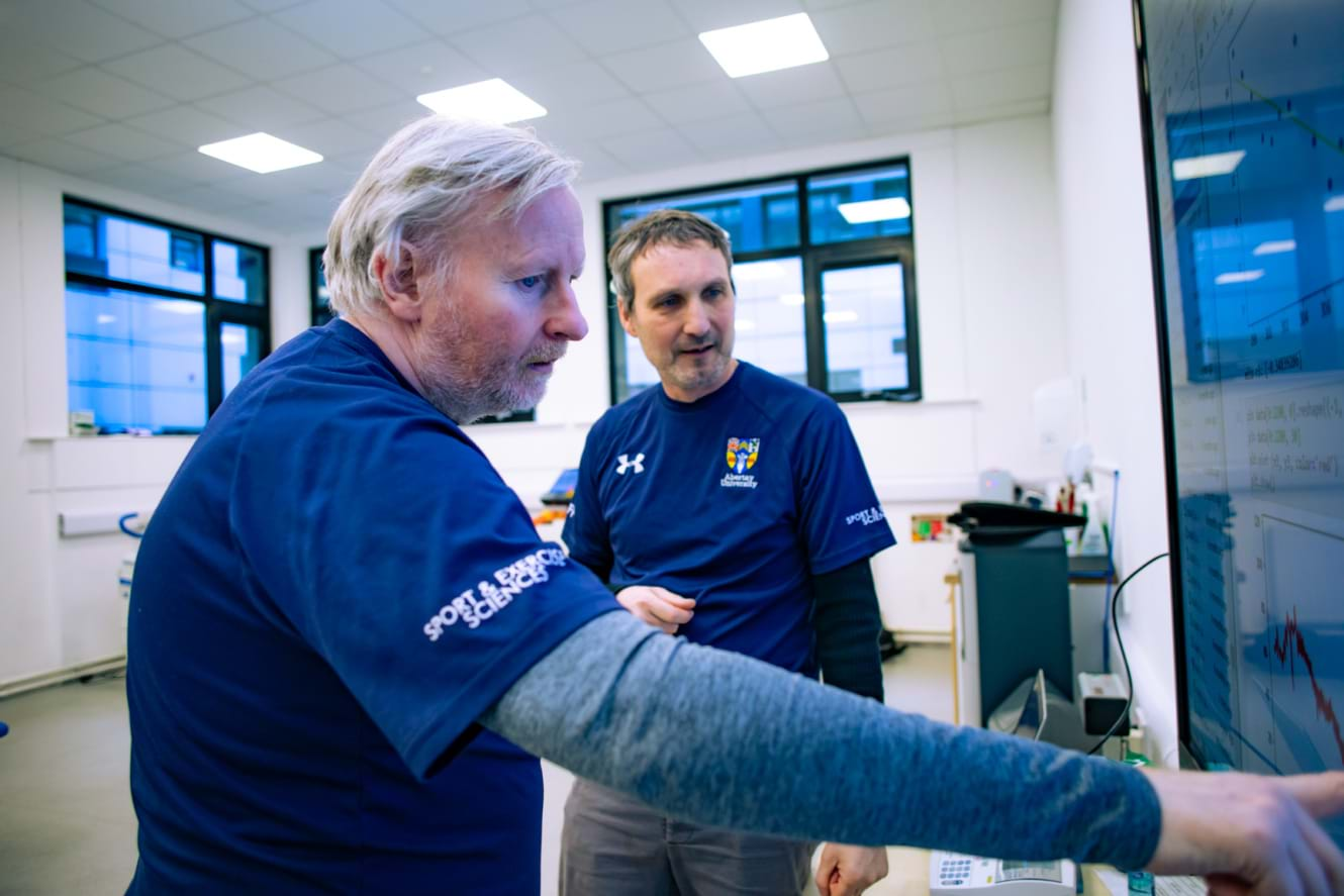 Two sports scientists working in a lab with a larger screen and equipment in the background