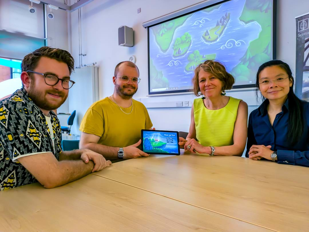 A group of four people sat around a table holding a tablet computer in the centre of the group.