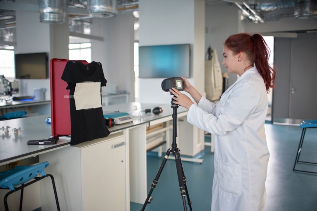 A woman in a white lab coat standing behind a camera, in front of her is a lab with a desk where a black t-shirt with paper attached that she is photographing. 