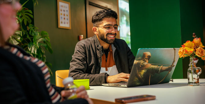 Male with short dark hair and glasses smiling while sitting in front of a laptop at a cafe table