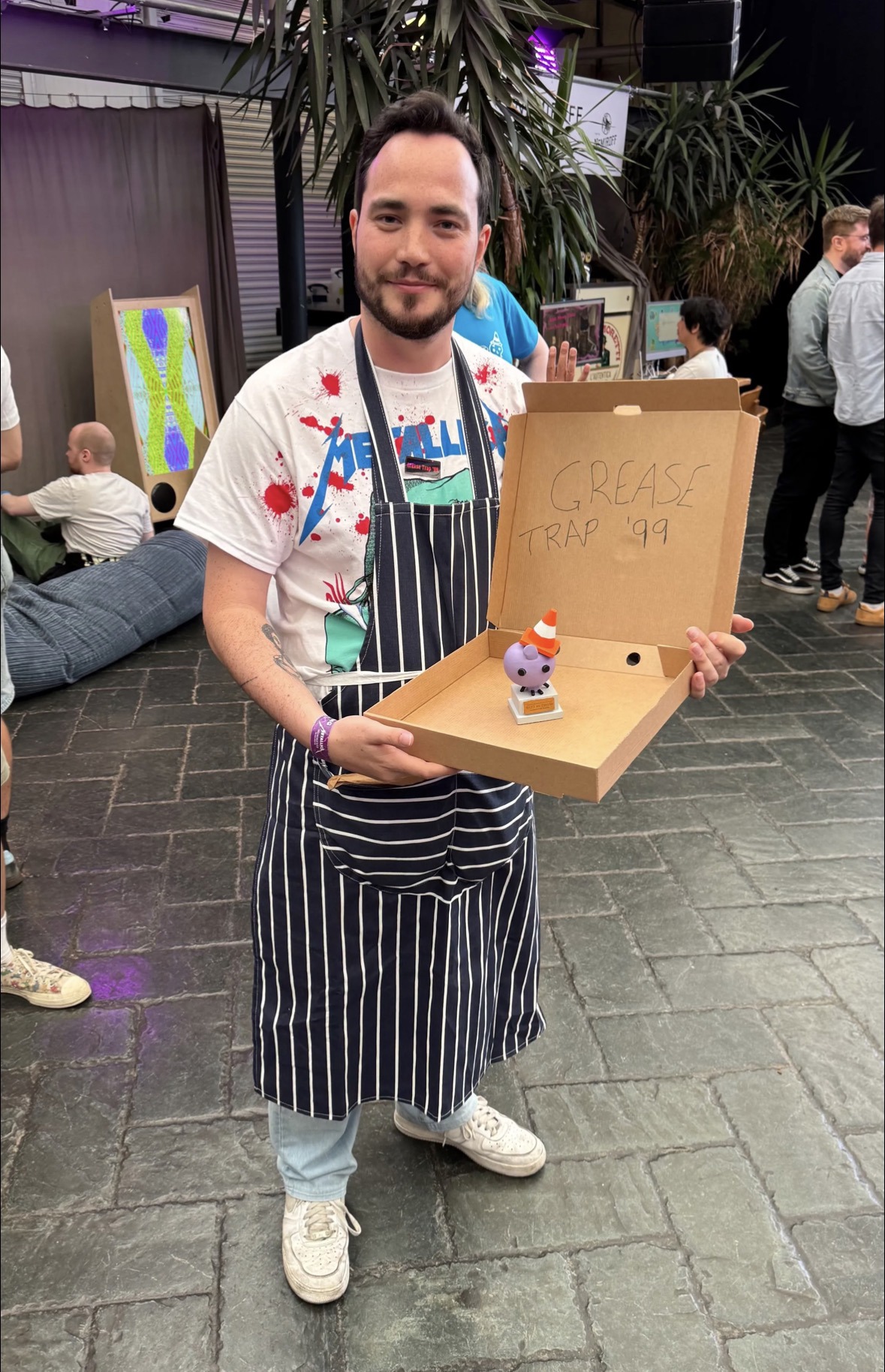 Abertay graduate James Muirhead at the Glasgow Indie Games Festival. He is dressed in a chip shop uniform in reference to his game, Grease Trap '99. He holds an open pizza box with a small figurine inside it.