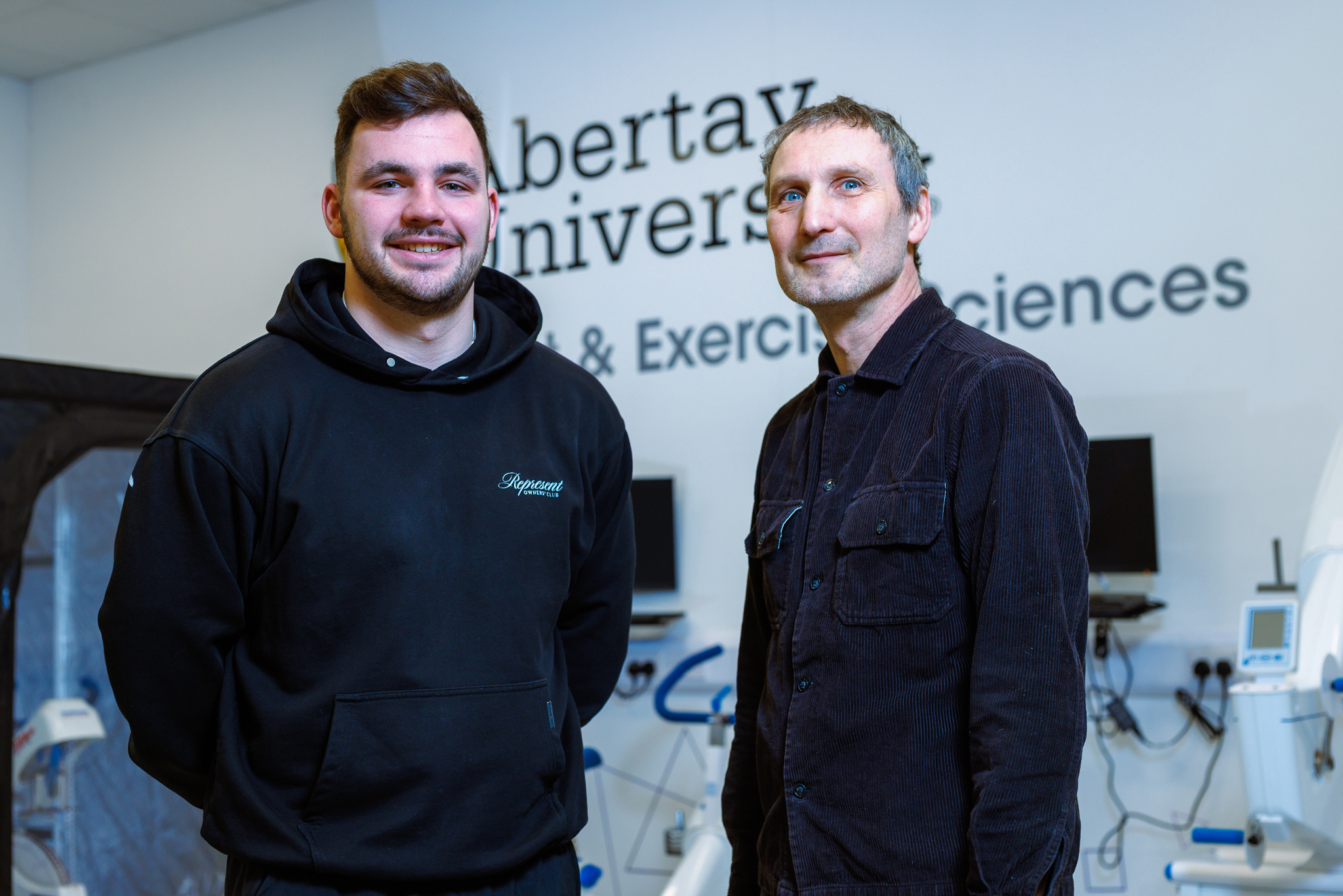 In the white-walled Abertay Human Performance Laboratory, student Adam Younger, smiling in a black hoody, stands on the left with his hands behind his back. To his right Dr John Babraj, in a black shirt, smiles to the camera.