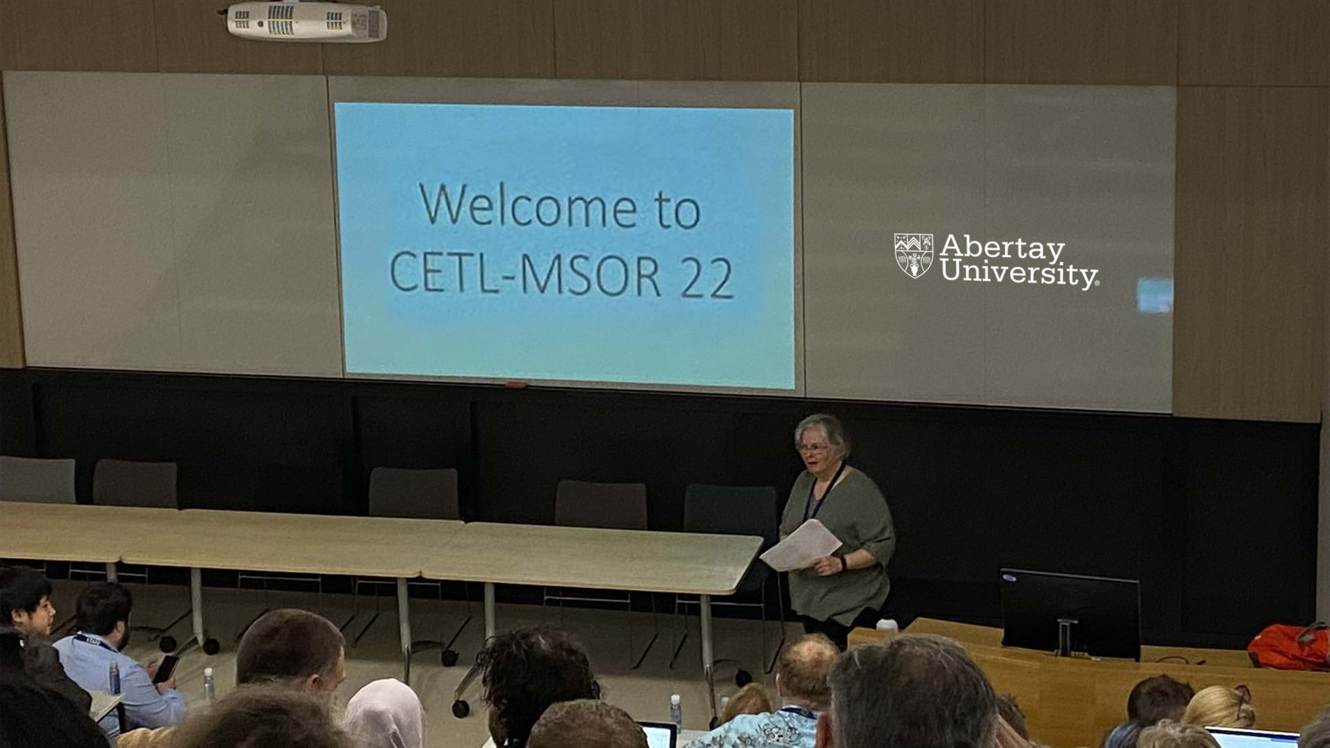 Conference main shot of a person presenting. In the background, a welcome slide is projected on the wall.